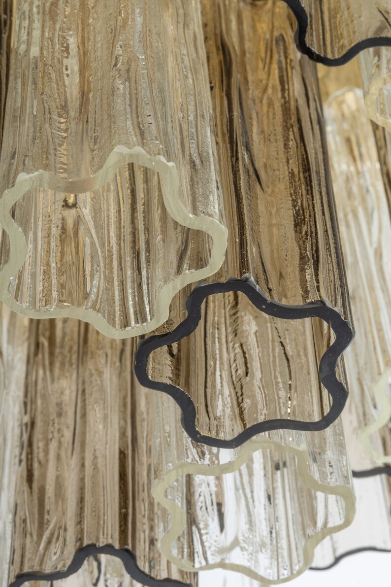 May include: Close-up of a glass chandelier with multiple tiers of clear glass tubes. The tubes have a textured, rippled surface and are joined together with black metal rings.