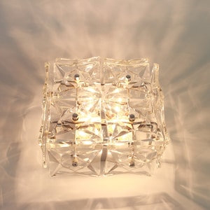 Könnte beinhalten: Eine transparente Glas-Deckenleuchte mit geometrischem Design. Die Leuchte hat eine quadratische Form und besteht aus mehreren kleinen, facettierten Glasscheiben. Die Leuchte wird mit warmweißem Licht beleuchtet.
