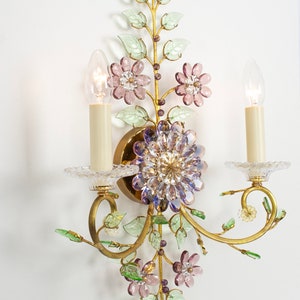 Puede incluir: Una l&aacute;mpara de pared dorada con un dise&ntilde;o floral. La l&aacute;mpara tiene dos bombillas de estilo vela y est&aacute; decorada con hojas verdes y flores rosas y moradas.