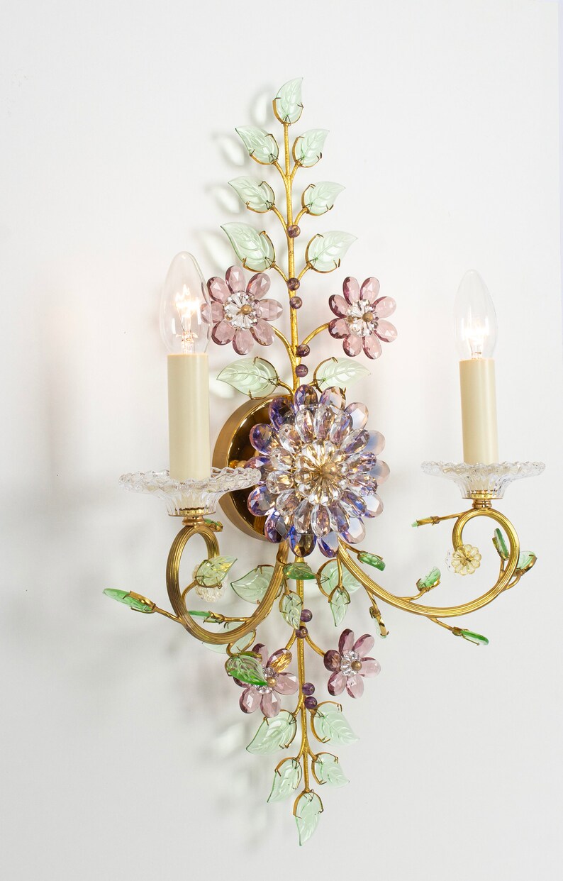 Puede incluir: Una l&aacute;mpara de pared dorada con un dise&ntilde;o floral. La l&aacute;mpara tiene dos bombillas de estilo vela y est&aacute; decorada con hojas verdes y flores rosas y moradas.
