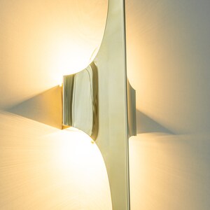 May include: A gold-toned wall sconce with a unique, curved design. The light fixture is mounted on a white wall and casts a warm glow.