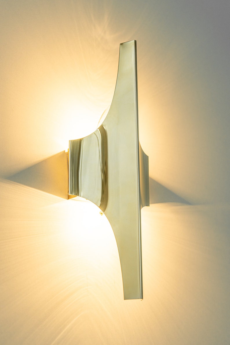 May include: A gold-toned wall sconce with a unique, curved design. The light fixture is mounted on a white wall and casts a warm glow.