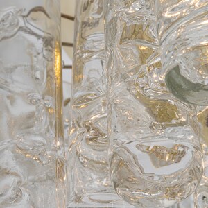 May include: A close-up of a chandelier with clear glass cylinders. The glass has a textured, bubbly appearance, and the light shines through the glass, creating a warm glow.