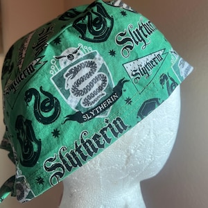 May include: A green scrub cap featuring a repeating pattern of black Slytherin house crests, snake icons, and pennants with the text Slytherin printed in a decorative font. The fabric has a vibrant green background with black graphic elements.