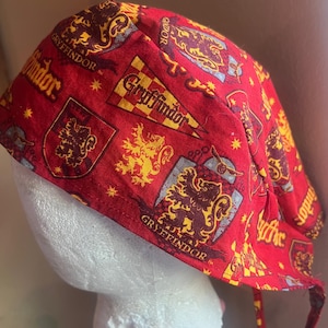 May include: A red fabric scrub cap featuring a repeating pattern of Gryffindor house crests, pennants, and lion emblems in yellow and dark red. The design includes the text Gryffindor printed in a bold yellow font throughout the fabric surface.