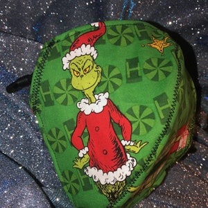 May include: Green fabric cube featuring a cartoon Grinch character in a red Santa suit and hat with white trim. The cube has a black drawstring and is decorated with green and white Christmas-themed patterns.