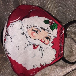 May include: Red and white face mask featuring a Santa Claus design. The mask has a red border and a white beard and face. The Santa Claus face has a jolly expression. Black elastic ear loops are attached.