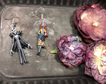 May include: Earrings featuring Jack Skellington and Sally from The Nightmare Before Christmas. Jack is in a black and white pinstripe suit, and Sally is in a patchwork dress. The earrings are displayed with purple fabric flowers.