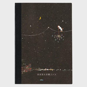 Könnte beinhalten: Notizbuch mit schwarzem Rücken und dunklem Einband mit einer skurrilen Illustration. Eine weiße Katze balanciert auf einem Drahtseil vor einem sternenklaren Nachthimmel, mit einem Mond und Stadtlichtern darunter. Japanischer Text unten.