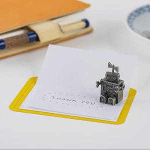 May include: A small silver-toned metal robot figurine with the words YOU LOVE YOU on its chest, standing on a white folded card with the text THANK YOU printed in black ink, framed by a yellow border, with a notebook and pen nearby.