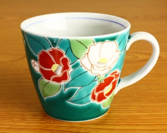 Puede incluir: Una taza de cerámica de color turquesa con un borde y un asa blancos. La taza está decorada con un patrón floral de camelias rojas, blancas y amarillas sobre un fondo de hojas verdes.
