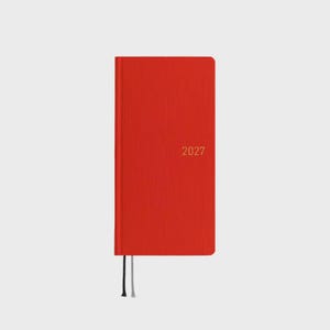Può includere: Un'agenda rettangolare sottile con una copertina rossa testurizzata. L'anno 2027 è stampato in un piccolo carattere color oro sulla copertina anteriore. Due sottili nastri segnalibro, uno nero e uno grigio chiaro, sporgono dal bordo inferiore.
