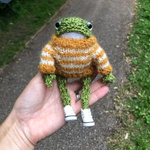 May include: A small, handmade green crochet frog toy featuring black plastic eyes and a white mouth area. The frog wears a knitted mustard yellow and white striped jumper with long sleeves and is wearing miniature white lace-up trainers.
