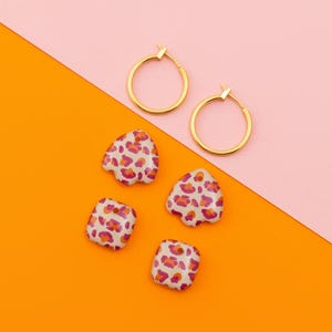 Puede incluir: Pendientes de aro dorados y cuatro piezas de joyería con estampado de leopardo sobre un fondo rosa y naranja. Las piezas de joyería tienen una base blanca con manchas rojas y naranjas. Los pendientes son circulares.