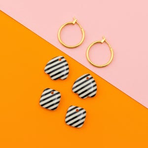 May include: Gold hoop earrings and four black and white striped charms in various shapes are arranged on a pink and orange background. The charms have small holes for hanging. The brand name "LA BELLE CAMILLE" is at the bottom.