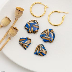 Peut inclure: Une composition de bijoux et d'éléments décoratifs sur une assiette blanche. L'image présente des créoles dorées et des pendentifs en forme de cœur avec des motifs de feuilles bleues et dorées. Des têtes de pavot séchées sont également présentes.
