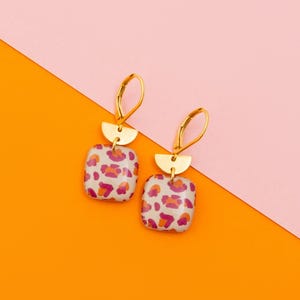 Puede incluir: Pendientes dorados con colgantes cuadrados estampados con diseños florales rojos y rosas sobre fondo blanco. Cada pendiente tiene un acento de semicírculo dorado y un cierre de palanca. El fondo está dividido diagonalmente, con secciones naranjas y rosas.