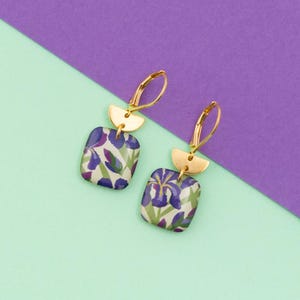 Peut inclure: Boucles d'oreilles dorées avec des pendentifs carrés à motifs floraux violets et verts. Chaque boucle d'oreille comporte un détail doré en demi-lune au-dessus du carré et un fermoir à levier. Le fond est divisé en diagonale, avec des sections violettes et vert menthe.