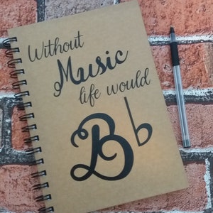 May include: A brown spiral bound notebook with the text "Without Music life would be" and a musical note and the letter "B" in black. The notebook is on a brick background.