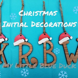 May include: Four wooden Christmas decorations shaped like the letters K, R, B, and W. Each letter is painted in a light brown colour and has a red and white Santa hat on top. The decorations are hanging from twine string against a turquoise background.