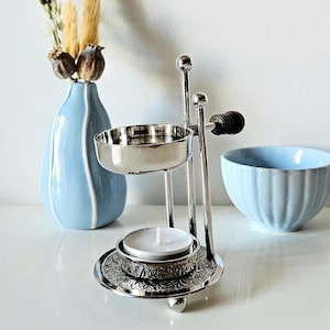 Pode incluir: Um queimador de óleo de aromaterapia prateado com uma vela, um vaso azul claro com flores secas e uma tigela azul clara. O queimador de óleo tem uma base decorativa e um prato pequeno para o óleo. O vaso e a tigela são de cerâmica.