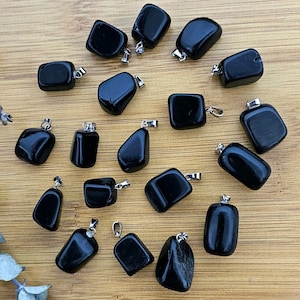 Könnte beinhalten: Eine Ansammlung von polierten schwarzen Obsidian-Anhängern mit silberfarbenen Ösen, präsentiert auf einer hellbraunen Holzoberfläche. Die Anhänger haben unterschiedliche Formen. Ein Zweig getrockneter grüner Blätter liegt links.