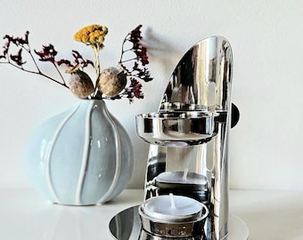 Könnte beinhalten: Ein polierter silberner Kerzenhalter mit zwei Ebenen für Teelichter. Links steht eine kleine, hellblaue Vase mit Trockenblumen. Der Kerzenhalter hat ein modernes, minimalistisches Design.