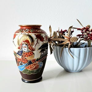 Könnte beinhalten: Eine dekorative japanische Vase mit einem detaillierten, bemalten Design mit einer Figur in traditioneller Kleidung. Die Vase ist braun, rot und goldfarben. Daneben eine hellblaue, gerippte Schale mit getrockneten Blumen.