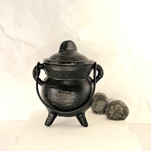May include: A small, black cast iron cauldron with a lid, two handles, and three legs. Two small, gray stones with carved designs of a Buddha and a four-leaf clover are next to the cauldron. The cauldron is on a white surface.