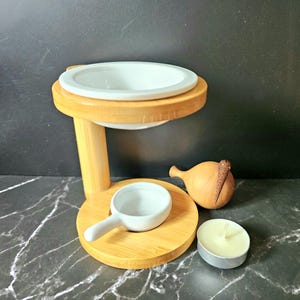Op de afbeelding: Een essentiële olie diffuser van bamboe en keramiek, met een witte keramische kom en een kleine witte keramische lepel. Een houten decoratie-item en een waxinelichtje zijn ook aanwezig.