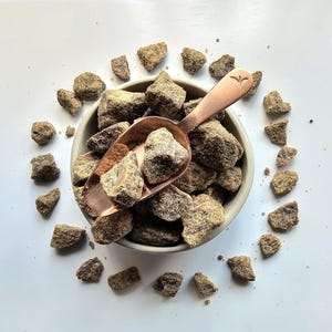 Könnte beinhalten: Nahaufnahme einer hellgrauen Schüssel gefüllt mit braunen Zuckerbrocken. Eine Kupferschaufel liegt in der Schüssel, ebenfalls gefüllt mit Zucker. Braune Zuckerstücke sind um die Schüssel auf einer weißen Oberfläche verstreut.