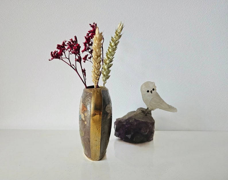 May include: A small, decorative vase with a brass and brown finish, holding dried red and tan flowers. A small, white owl figurine sits on a purple amethyst crystal, creating a natural, artistic display.