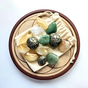 Pu&ograve; includere: Una collezione di pietre preziose lucide di vari colori, tra cui verde, giallo e trasparente, disposte su un sottobicchiere di legno con un design geometrico. Le pietre sono in un piccolo sacchetto di cordoncino color crema.