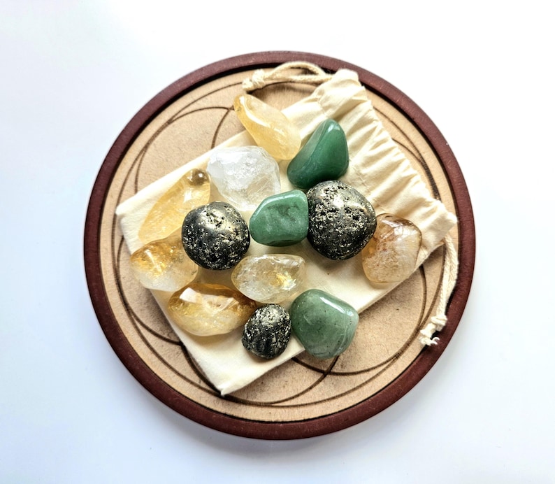 Pu&ograve; includere: Una collezione di pietre preziose lucide di vari colori, tra cui verde, giallo e trasparente, disposte su un sottobicchiere di legno con un design geometrico. Le pietre sono in un piccolo sacchetto di cordoncino color crema.