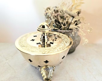 May include: A gold-colored incense burner with a domed lid and decorative carvings. The burner has a round base with three ornate feet and a central handle. The lid features small cut-out shapes. A vase of dried flowers is in the background.