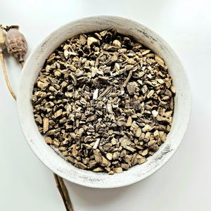 Puede incluir: Un cuenco de cerámica blanco desgastado lleno de pequeños trozos picados de hierbas secas de color marrón claro. Dos vainas de semillas secas están colocadas a la izquierda del cuenco. El fondo es una superficie blanca brillante.