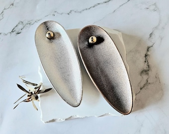 May include: Two metallic, oval-shaped decorative dishes. One is silver, the other is bronze. Each has a small, gold-coloured sphere at one end. The dishes rest on a white, textured surface, with a dried flower nearby.