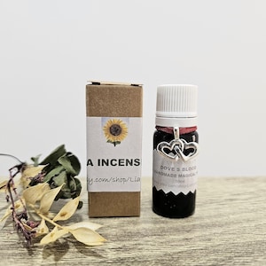 Op de afbeelding: Een zwarte glazen fles met een witte dop en een zilveren bedel met twee harten. Naast de fles staat een kleine bruine doos met een zonnebloemafbeelding en de tekst "A Incens". Gedroogde bloemen en bladeren staan op de voorgrond.