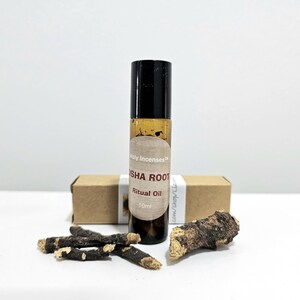 Può includere: Una piccola bottiglia di vetro scuro con tappo nero, etichettata "OSHA ROOT Ritual Oil 10ml", è posizionata accanto a una piccola scatola di cartone marrone e diversi pezzi di radice essiccata. La bottiglia contiene un liquido dorato. Gli oggetti sono disposti su una superficie bianca.