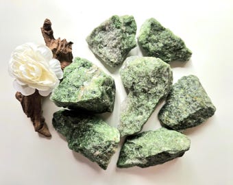 May include: A collection of rough, green stones with a textured surface. A white flower and a piece of driftwood are arranged near the stones. The stones vary in size and shape, with a natural, earthy appearance.
