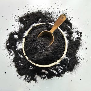 Op de afbeelding: Een ronde witte kom gevuld met fijn zwart poeder, omringd door een verspreide hoop van dezelfde donkere substantie. Een kleine goudkleurige metalen schep met een bladvormig embleem op het handvat ligt in de kom, deels begraven in het poeder.