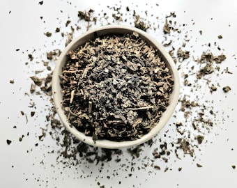 May include: Overhead shot of a small, light-colored ceramic bowl filled with dried, chopped green herbs. The herbs are dark green and brown, with some scattered around the bowl on a white surface. The image is well-lit and the focus is sharp.