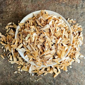 Puede incluir: Un cuenco blanco lleno de trozos de madera triturada de color marrón claro. El cuenco se encuentra sobre una superficie marrón texturizada. Las virutas de madera se derraman del cuenco, creando una estética natural y terrosa. La imagen está tomada desde arriba.