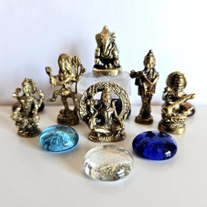 Puede incluir: Una colección de pequeñas estatuas doradas de deidades hindúes, incluyendo a Ganesha y Shiva, dispuestas con piedras de vidrio azules y transparentes. Las estatuas son detalladas sobre un fondo blanco.