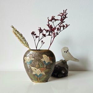 May include: A decorative vase with a floral design in shades of brown, blue, and orange, holding dried flowers and wheat. A small, white owl figurine sits on a dark rock, adding a touch of nature-inspired decor.