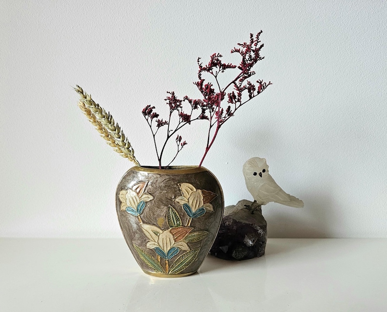May include: A decorative vase with a floral design in shades of brown, blue, and orange, holding dried flowers and wheat. A small, white owl figurine sits on a dark rock, adding a touch of nature-inspired decor.