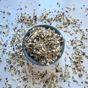 Könnte beinhalten: Eine dunkelgraue Schale gefüllt mit getrockneten, gehackten botanischen Wurzelstücken in Beige-, Braun- und Weißtönen. Die Schale ist zentriert, mit zusätzlichen losen, fragmentierten Pflanzenteilen, die um die Basis auf einer weißen Fläche liegen.