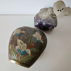 May include: A decorative, dark brown, metal container with a floral design in white, blue, and brown. The container has a gold-colored rim. A purple crystal and a white owl figurine are in the background.