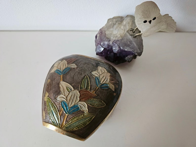 May include: A decorative, dark brown, metal container with a floral design in white, blue, and brown. The container has a gold-colored rim. A purple crystal and a white owl figurine are in the background.