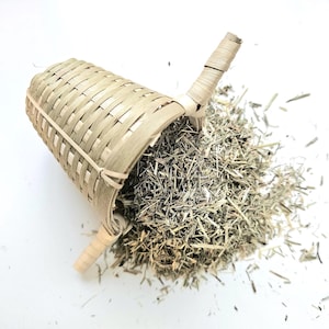 Könnte beinhalten: Ein gewebter Bambus-Teesieb, zur Seite geneigt, mit getrockneten Teeblättern, die herausfallen. Das Sieb hat eine gebogene Form mit einem Griff auf jeder Seite. Die Teeblätter sind eine Mischung aus Grün und Braun.